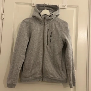 Grå huvtröja från Sail Racing - Säljer en stilren grå huvtröja från Sail Racing med dragkedja framtill och en praktisk bröstficka med dragkedja. Tröjan har en bekväm huva med dragsko och långa ärmar. Perfekt för en avslappnad look.