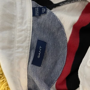 Randig pikétröja från GANT - Säljer en snygg randig pikétröja från GANT i färgerna röd, svart och vit. Tröjan har långa ärmar och texten 'EAST COAST NEW HAVEN' på kragen. Perfekt för en avslappnad stil.