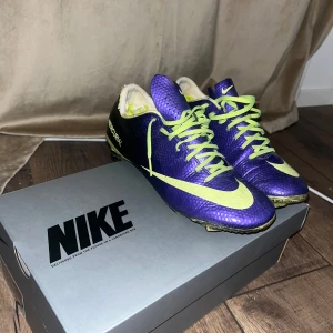 Lila och gula Nike fotbollsskor storlek 41/26cm - Säljer ett par lila och gula Nike fotbollsskor med snörning. Skorna har en sportig design med en iögonfallande färgkombination och är perfekta för fotbollsplanen. De har en slitstark sula med dobbar för bra grepp.