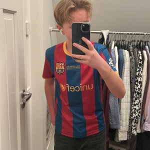 Blåröd fotbollströja från FC Barcelona - Säljer en blåröd randig fotbollströja från FC Barcelona med Neymar Jr och nummer 11 på ryggen. Tröjan har korta ärmar med gula detaljer och ett tryck av Champions League-loggan på ärmen. Perfekt för fans av laget!