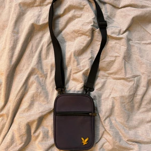 Svart axelväska från Lyle&Scott - Snygg och praktisk svart axelväska med justerbar rem och dragkedja. Väskan är perfekt för att bära med sig det nödvändigaste. Enkel och stilren design som passar till många tillfällen.