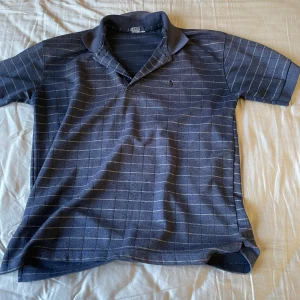 Blå pikétröja från Ralph Lauren - Säljer en stilren blå pikétröja från Ralph Lauren med diskret rutmönster och broderad logga på bröstet. Tröjan är tillverkad i 100% bomull och har korta ärmar. Perfekt för en avslappnad men ändå snygg look.