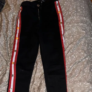 Svarta högmidjade jeans med röda ränder - Snygga svarta högmidjade jeans från Vice med röda och vita ränder längs sidorna. Byxorna har dekorativa knappar i guld och silver på ränderna och en dragkedja framtill. Perfekta för en trendig look! Använd 1 gång.