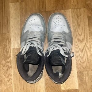 Nike Air Jordans i grått och vitt - Säljer ett par klassiska Nike Air Jordans i grått och vitt med svart swoosh. Skorna har snörning och perforerade detaljer på tån för extra ventilation. Perfekta för dig som älskar en sportig stil.