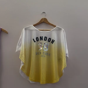Vit och gul topp med paljetter - Säljer en vit och gul topp med texten 'London 36 New York Tokyo' i svart och paljetter. Toppen har en lös passform och korta ärmar, perfekt för en avslappnad stil. storlek 14+