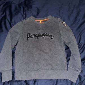 Grå fleece tröja från Parajumpers - Säljer en grå fleece tröja från Parajumpers med svart text på framsidan och en patch på ärmen. Tröjan har långa ärmar och orange detaljer vid halsen. Perfekt för kyliga dagar!