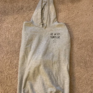 Grå hoodie från Gina Tricot - Säljer en grå hoodie från Gina Tricot med texten 'Je m'en amour' och en stjärna tryckt på framsidan. Hoodien har en klassisk design med huva och ribbade muddar vid ärmslut och nederkant. Perfekt för en avslappnad stil.