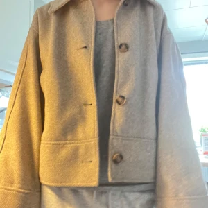 Beige kappa med knappar - Säljer en stilren beige kappa med knappar framtill. Jackan har en klassisk krage och långa ärmar. Perfekt för höst och vår, tillverkad i ett mjukt material som ger en bekväm känsla!!