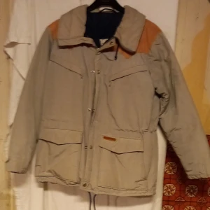 Beige dunjacka från Tenson - Säljer en beige dunjacka från Tenson med bruna axelpartier. Jackan har långa ärmar och flera praktiska fickor framtill. Den är tillverkad i ett slitstarkt material och har en dragkedja samt knappar för stängning. Perfekt för kyligare väder!