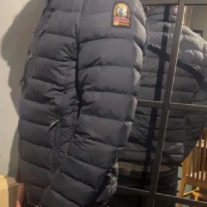Mörkblå dunjacka från Parajumpers - Säljer en snygg mörkblå dunjacka från Parajumpers. Jackan har en dragkedja framtill och en hög krage med en gul detalj. Den är quiltad och har långa ärmar samt en logotyp på ärmen. Perfekt för kyligare dagar!