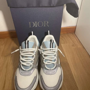 Sneakers från Dior - Snygga sneakers från Dior i en stilren design med vita och gråa nyanser. Skorna har snörning och en modern look som passar perfekt för en trendig stil. De kommer i originalförpackning med Dior-logga.