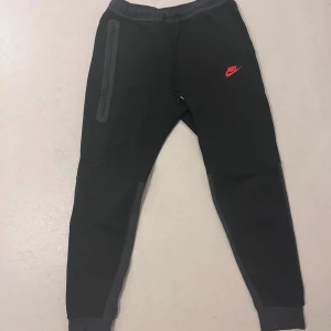 Nike tech byxor - Snygga svarta mjukisbyxor från Nike med en röd logga på framsidan. Byxorna har en dragkedja på sidan och elastisk midja för extra komfort. Perfekta för en avslappnad stil.