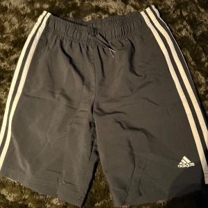 Svarta träningsshorts från Adidas - Snygga svarta träningsshorts från Adidas med klassiska vita ränder längs sidorna. Shortsen har elastisk midja med dragsko för bästa passform. Perfekta för sport och träning.