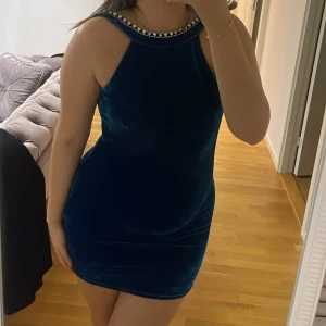 Blå sammet klänning med pärldetaljer - Snygg blå sammet klänning med pärldetaljer runt halsringningen och ärmlös design. Klänningen har en tight passform och en öppen rygg som ger en elegant touch. Perfekt för en kväll ute eller speciella tillfällen.