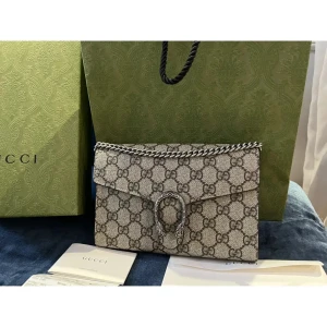 Äkta Gucci kuvertväska med kedjedetalj - Säljer en elegant Gucci kuvertväska med det ikoniska GG-mönstret. Väskan har en stilren kedjedetalj och flera fack inuti för smidig organisering. Perfekt för att ge en lyxig touch till din outfit. Kommer med originalförpackning och certifikat.