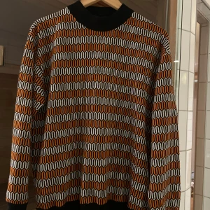 Mönstrad tröja från Pasaco - Säljer en unik och stilren tröja från det franska märket Pasaco med ett coolt vågmönster i svart, vitt och orange. Tröjan är i gott skick och har bekväm passform. 