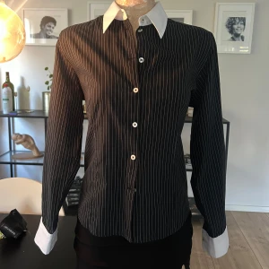  Randig figursydd skjorta  - Säljer en stilren svart skjorta med vita ränder och kontrasterande vita krage och manschetter. Skjortan har en slim passform och långa ärmar med knappar fram!  Storlek 38 på lappen men passar mer 34/36