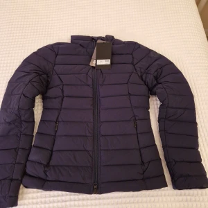 Mörkblå dunjacka från J.Lindeberg - Snygg och varm mörkblå dunjacka från J.Lindeberg. Jackan har en stilren design med dragkedja framtill och långa ärmar. Perfekt för kyliga höst- och vinterdagar. Den är helt ny med alla lappar kvar.