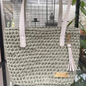 Stickad väska med tofsdetalj - Säljer en superfin virkad väska i en mjuk grön nyans. Den har vita handtag och en snygg tofsdetalj i vitt och guld. Väskan är perfekt för både vardag och speciella tillfällen. Den har en stabil botten med små metallfötter för att skydda mot slitage.