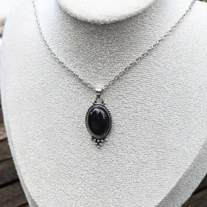 Gothic halsband med färgskiftande sten - Elegant gothic halsband med en stor, oval svart sten i mitten. Hänget har en detaljerad kant med små silverkulo.alrig använt  Det är svårt att fånga de exakta färgerna, men stenens nyans skiftar vackert mellan brunt ,svart och rött beroende på ljuset