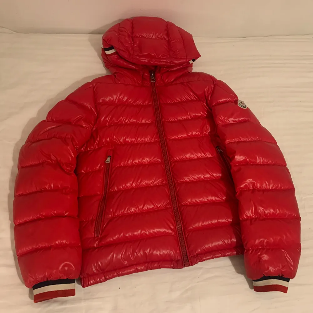 Säljer en snygg röd dunjacka från Moncler. Jackan har en quiltad design med en dragkedja framtill och en huva. Den är perfekt för kalla dagar och har långa ärmar med ribbade muddar i rött, vitt och blått. PRIS KAN DISKUTERAS. Jackor.