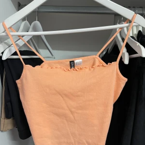 Persikofärgad croppad linnetopp från Divided - Säljer en superfin persikofärgad croppad linnetopp från Divided. Den har smala axelband och en söt volangdetalj längs kanten. Perfekt för sommardagar eller en avslappnad festkväll. Toppen är i storlek L, passar M, oanvänd!