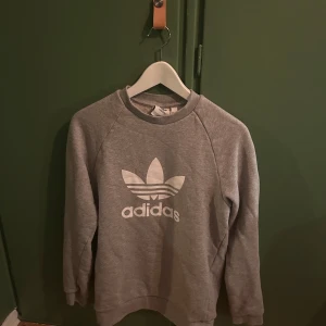 Grå sweatshirt från Adidas - Säljer en grå sweatshirt från Adidas i mycket bra skick. Den har det klassiska Adidas-loggan tryckt på framsidan. Tröjan är långärmad och perfekt för både vardag och träning. Superbekväm och stilren design! Obs! Ett litet hål i ena armhålan. Men det är ej synligt när tröjan är på!