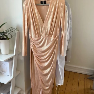 Champagnefärgad / rosa / guld långklänning med drapering och slits - Säljer en elegant champagnefärgad långklänning i glittrigt material. Klänningen har en v-ringad design och långa ärmar, perfekt för festliga tillfällen. Den har en vacker drapering framtill som ger en smickrande passform. Perfekt för nyår eller andra speciella tillfällen! 