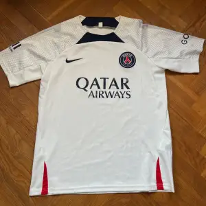 Säljer en vit PSG fotbollströja från Nike i mycket bra skick. Den har korta ärmar och snygga detaljer i mörkblått och rött. Perfekt för alla fans av Paris Saint-Germain! Tröjan har Qatar Airways som sponsorlogga på framsidan. Passar perfekt för match eller träning. Hör av dig vid frågor👋