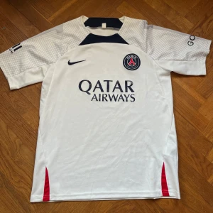  PSG fotbollströja - Säljer en vit PSG fotbollströja från Nike i mycket bra skick. Den har korta ärmar och snygga detaljer i mörkblått och rött. Perfekt för alla fans av Paris Saint-Germain! Tröjan har Qatar Airways som sponsorlogga på framsidan. Fick i present förra året så lite osäker på autencitet. Passar perfekt för match eller träning. Hör av dig vid frågor👋
