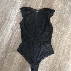 Svart spetsbody - Snygg svart body med spetsdetaljer och korta ärmar. Perfekt för en festlig kväll eller som en del av en lager-på-lager-look. Den har en elegant design med spets som ger en lyxig känsla.