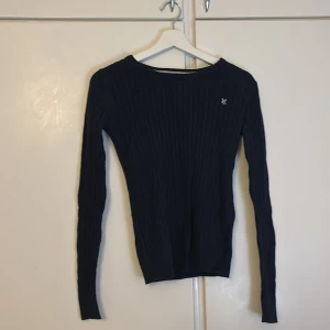 Mörkblå ribbad tröja - Säljer en snygg mörkblå ribbad tröja med ett diskret broderat märke på bröstet. Tröjan har en klassisk passform med långa ärmar och är perfekt för både höst och vinter. Den är i mycket bra skick och passar till alla tillfällen. Perfekt för dig som vill ha en stilren look! 💙