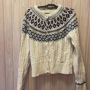 Vit stickad kofta från Lindex - Säljer en superfin vit stickad kofta från Lindex med ett snyggt mönster i blått och rosa upptill. Koftan har långa ärmar och knappar framtill. Perfekt för kyliga höst- och vinterdagar. Den är i mycket bra skick och redo för en ny ägare!