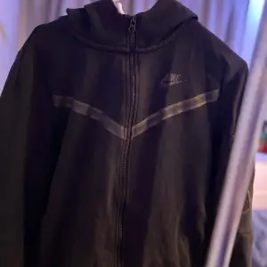 Säljer en svart hoodie från Nike i bra skick. Den har en dragkedja framtill och en praktisk ficka med dragkedja på ärmen. Perfekt för höst och vår, med en stilren design och Nike-logga på bröstet. Passar både till träning och vardag. 