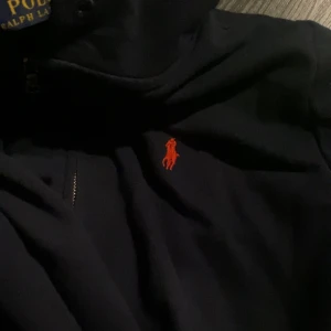 Mörkblå hoodie från Ralph Lauren - Säljer en snygg blå hoodie från Ralph Lauren med det klassiska röda logotypen på bröstet. Den har en dragkedja framtill och är perfekt för både höst och vår. Passar bra till vardags och är superbekväm! Obs priset är inte fast i sten:)