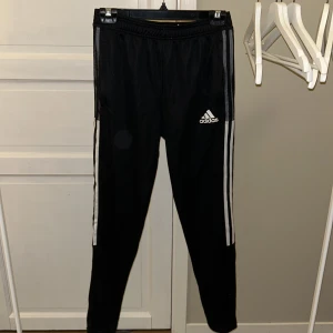 Svarta träningsbyxor från Adidas - Säljer ett par svarta träningsbyxor från Adidas i mycket bra skick. De har de klassiska tre ränderna längs sidorna och är tillverkade i ett lätt och andningsbart material med AEROREADY-teknologi. Perfekta för träning eller en avslappnad dag. Storlek 13-14 år (164), men de ser ut att ha en normal passform.