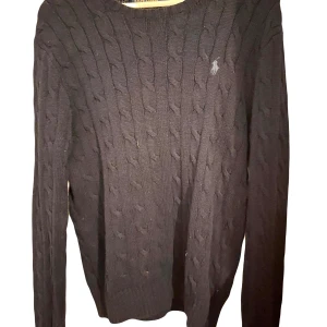 Svart stickad tröja från Polo Ralph Lauren - Säljer en snygg svart stickad tröja från Polo Ralph Lauren. Den har ett klassiskt kabelstickat mönster och en liten broderad logga på bröstet. Perfekt för både höst och vinter, med långa ärmar och ribbade kanter. Passar till både vardag och fest!