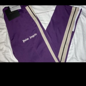 Lila träningsbyxor från Palm Angels - Snygga lila träningsbyxor från Palm Angels med vita och svarta ränder längs sidorna. Byxorna har en elastisk midja och en broderad logga på sidan. Perfekta för en sportig look.