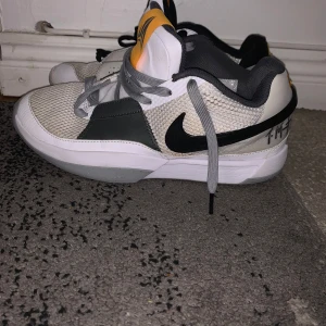 Ja 1 - Säljer ett par Nike sneakers med grå och vita detaljer samt en svart swoosh. Skorna har en unik design med olika färger på snörena och en orange detalj på plösen. Perfekta för en sportig look.