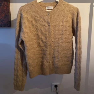 Beige stickad kofta från Holly & Whyte - Säljer en mysig beige stickad kofta från Holly & Whyte by Lindex. Koftan har ett fint flätmönster och knappar framtill. Perfekt för kyliga dagar!