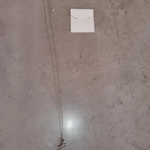 Halsband med skridskohänge - Snyggt halsband med ett unikt skridskohänge i silverfärg. Perfekt för den som älskar vintersport eller bara vill ha ett annorlunda smycke. Kedjan är tunn och elegant, vilket gör att hänget verkligen står ut.