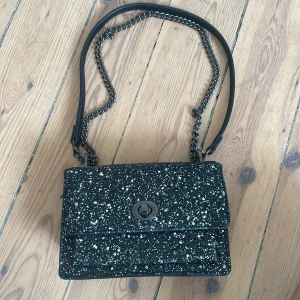 Glittrig väska  - Snygg svart axelväska med glittrigt mönster och kedjedetaljer. Väskan har en praktisk storlek med flera fack inuti och en vridlåsning framtill. Perfekt för att ge en extra touch till din outfit. Använd 1-3 gånger