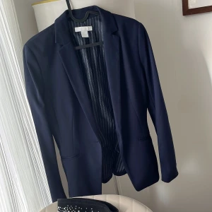 Mörkblå kavaj från H&M - Snygg mörkblå kavaj från H&M med ett stilrent randigt foder. Perfekt för en elegant look. Kavajen har en klassisk design med krage och är tillverkad i ett mjukt material som ger en bekväm passform.