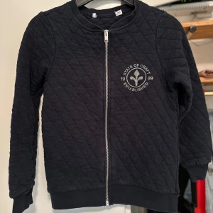 Svart quiltad jacka från State of Craft storlek: 150 - Säljer en svart quiltad jacka från State of Craft med dragkedja framtill. Jackan har ett tryck med texten 'State of Craft Established 1999' på bröstet. Perfekt för en stilren look.