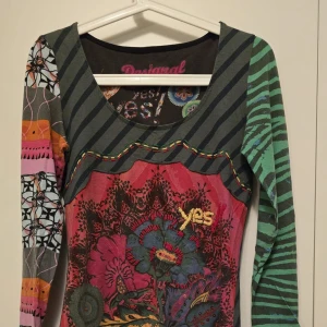 Mönstrad långärmad tröja från Desigual - Unik och färgglad långärmad tröja från Desigual med ett mixat mönster av ränder och blommor. Tröjan har en kombination av olika färger som grön, rosa och orange, vilket ger den en livfull look. Perfekt för dig som vill sticka ut!