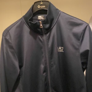 Mörkblå jacka från EA7 - Snygg mörkblå jacka från EA7 med dragkedja framtill. Jackan har en hög krage och långa ärmar, perfekt för en stilren look. Den är tillverkad i ett syntetmaterial som ger en modern känsla.