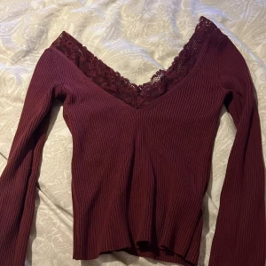 Vinröd ribbad topp från H&M Divided - Säljer en vinröd ribbad topp från H&M Divided med spetsdetaljer vid den v-ringade halsen. Toppen har långa ärmar och en figurnära passform. Perfekt för en stilren look. 💫