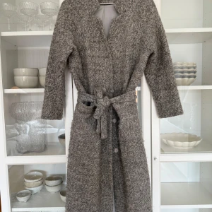Grå ullkappa från Gina Tricot - Snygg grå kappa i ullblandning från Gina Tricot. Den har en klassisk design med knappar och ett bälte i midjan för en elegant look. Perfekt för kyligare dagar med sin långa passform och mjuka material. Den har använts flitigt och har några skavanker, exempelvis har den gått upp lite i sidan (se bild) men det går enkelt att åtgärda. Öglan som skärpet ska sitta i saknas också på den sidan.