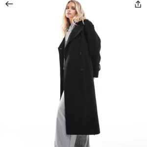 Svart lång kappa - Snygg svart lång kappa med dubbelknäppning och breda slag. Den har ett bälte i midjan och knappar vid ärmsluten. Perfekt för en stilren look.