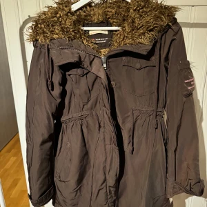 Brun parkajacka från Odd Molly - Säljer en brun parkajacka från Odd Molly med fuskpäls på huvan. Jackan har dragkedja och knappar framtill samt justerbar midja för en bättre passform. Perfekt för kyligare dagar.